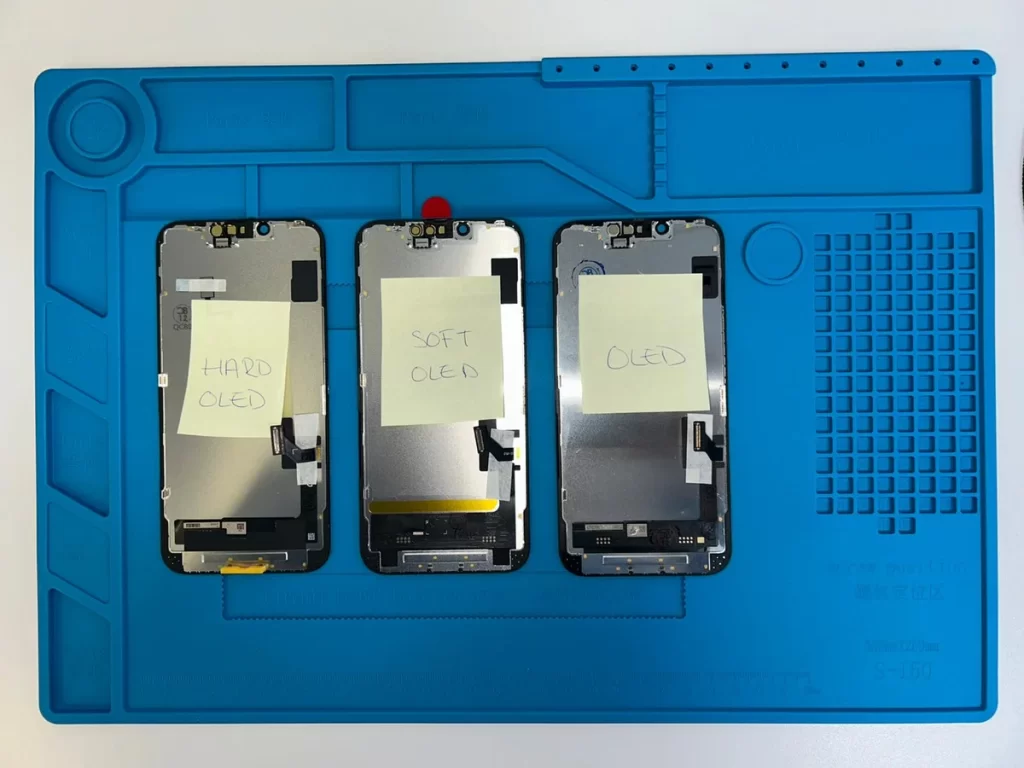 Tres pantallas para iPhone sobre tapete de reparación, etiquetadas como Hard OLED, Soft OLED y Excellent para comparar calidades.