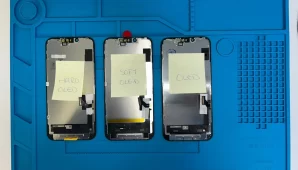 Tres pantallas para iPhone sobre tapete de reparación, etiquetadas como Hard OLED, Soft OLED y Excellent para comparar calidades.